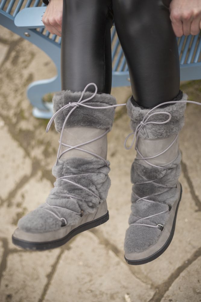 Nahaufnahme einer Person, die auf einer blauen Bank sitzt und schwarze Leggings und graue TORQ Winterstiefel von ZAQQ mit Kreuzschnürung trägt. Der Boden ist mit Steinen gepflastert und die Stiefel versprechen trotz ihrer robusten Bauweise ein gemütliches Barfußgefühl.