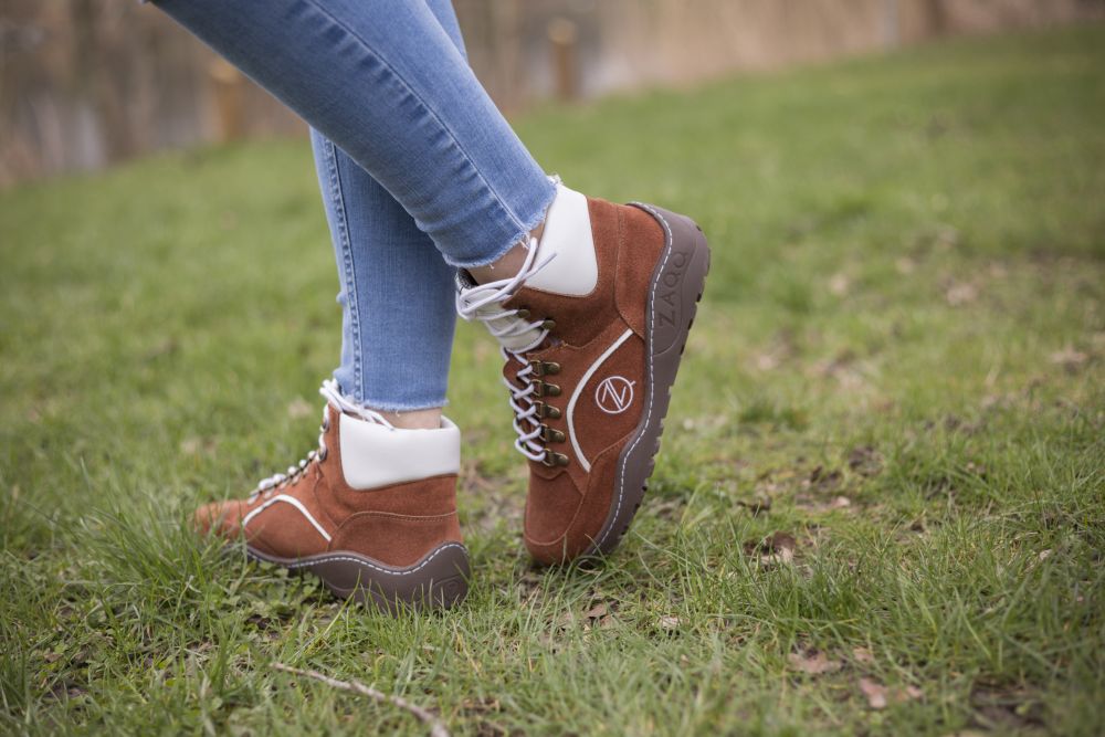 Eine Person steht auf Gras und trägt Blue Jeans und wasserdichte TREQ Cognac-Stiefel von ZAQQ, die sich durch ihre robusten Sohlen und weißen Akzente auszeichnen und darauf schließen lassen, dass sie für den Außenbereich konzipiert sind. Die Szene scheint in einer natürlichen Umgebung angesiedelt zu sein.