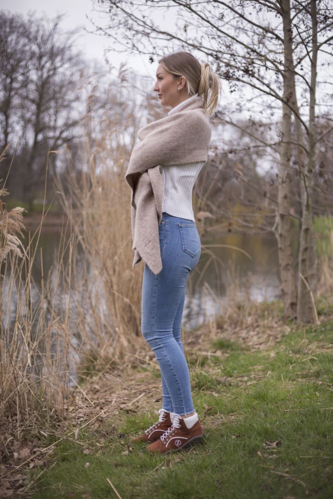 Eine Person steht auf einer Wiese an einem See, ist in einen beigen Schal gehüllt und trägt ein weißes Oberteil, blaue Jeans und braune wasserdichte Schuhe TREQ Cognac von ZAQQ. Sie blickt in die Ferne, mit kahlen Bäumen und getrocknetem Schilf im Hintergrund.