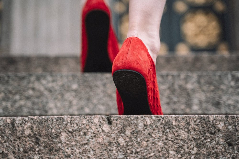 Nahaufnahme einer Person in ZAQQs TWIST Red High Heels, die Steintreppen hinaufgeht. Das leuchtende Velourleder kontrastiert mit den grauen Stufen und zeigt nur die Schuhe und Unterschenkel vor dem kunstvollen architektonischen Hintergrund.