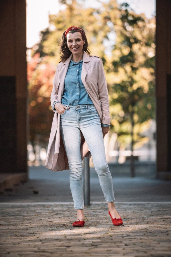 Eine Person steht draußen auf einem Steinweg, trägt hellblaue Jeans, ein Jeanshemd, einen rosa Trenchcoat und TWIST Velours Red Schuhe von ZAQQ mit einer glatten Kautschuksohle. Sie hat ein rotes Stirnband und lächelt mit einer Hand in der Tasche, während im Hintergrund Bäume und Gebäude verschwimmen.