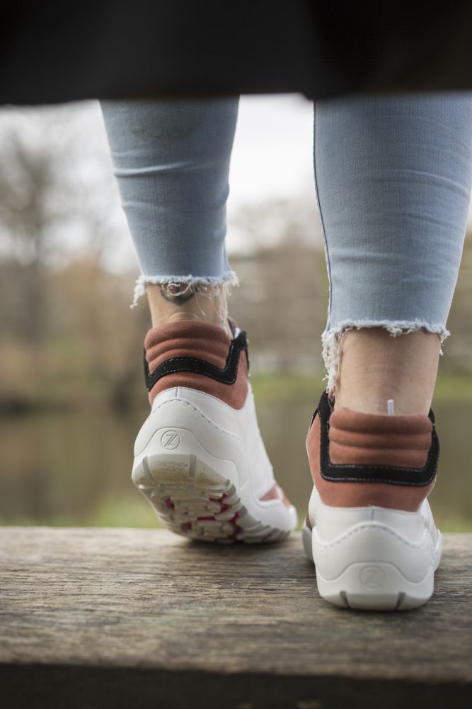 Eine Person in ausgefranster, hellblauer Jeans und weißen Sneakers mit pinken und schwarzen Akzenten steht auf einer Holzfläche. Die ZAQQ VAQATION Rosa Waterproof Schuhe sorgen für eine unvergleichliche Atmungsaktivität. Im Hintergrund ist ein verschwommener Blick auf einen Park mit Bäumen und einem Teich zu sehen.