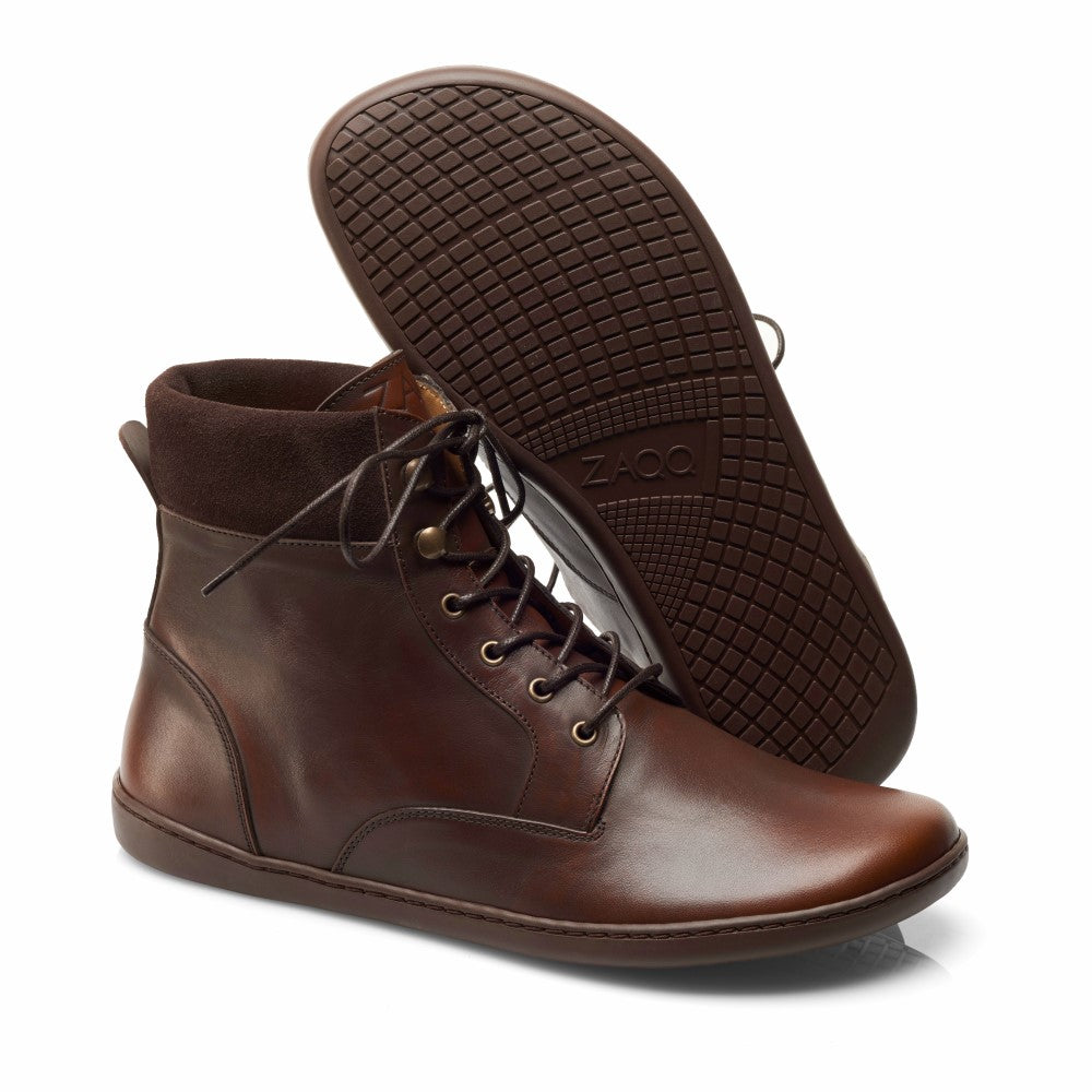 Ein Paar QORBY Brown High-Top-Schuhe von ZAQQ, gefertigt aus braunem Leder und mit passenden braunen Schnürsenkeln und Gummisohlen für optimalen Komfort. Ein Schuh steht aufrecht, während der andere angewinkelt ist, um das detaillierte Muster der Sohle zu zeigen.