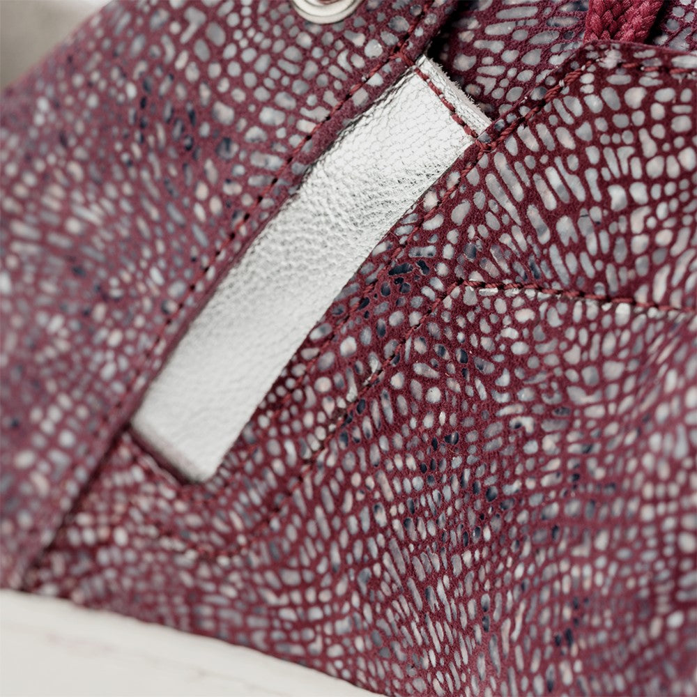 Nahaufnahme eines Sneakers mit roter und silberner Textur, der komplizierte Muster auf dem Stoff zeigt. Der ORQNEY Rosa von ZAQQ verfügt über einen kleinen silbernen Akzent, auffällige Nähte und Details aus Nappaleder, alles ergänzt durch die charakteristische ZAQQ-Kautschuksohle für ultimativen Komfort.