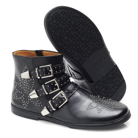 Die Stiefelette ZAQQ QLANCE Black Silver präsentiert sich in schwarzem Leder mit silbernen Nieten, dekorativen Schnallen und drei verstellbaren Riemen. Ein Stiefel steht aufrecht und zeigt das seitliche Design, während der andere eine gemusterte Sohle aus superweichem Leder aufweist.